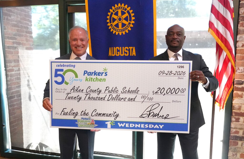 Two men in suits holding a large check reading "Parker's Kitchen" with "20,000" dollars. One man signs the check.