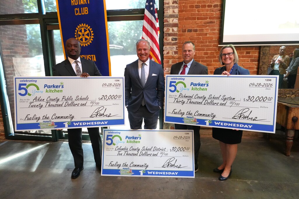 Four people hold large checks, with one individual standing behind the American flag. A brick wall is in the background.