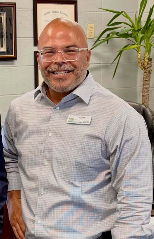 Man with a beard and glasses smiles. He wears a light blue button-down shirt with a name tag.