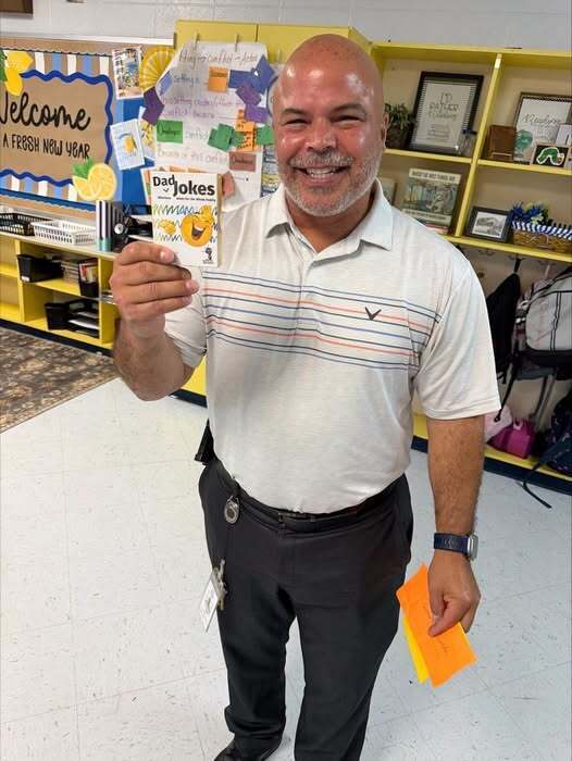 A smiling man holds a yellow card and a card reading "Dad Jokes" in a classroom with yellow shelves and a white floor.