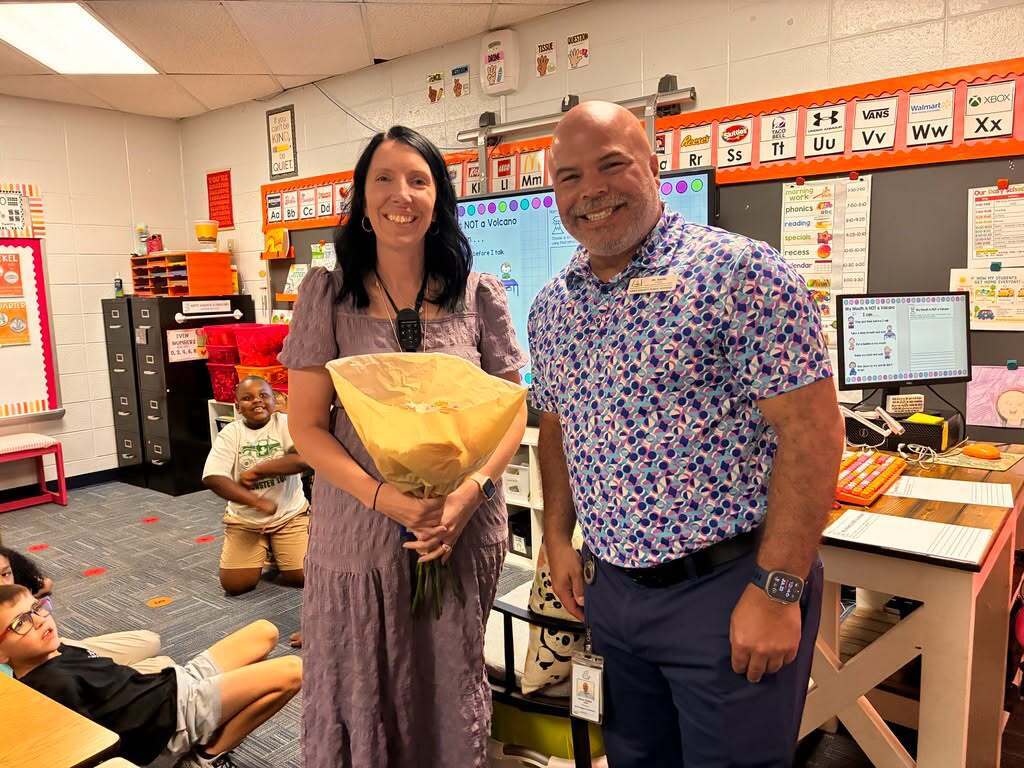 A woman and man standing in a classroom, holding flowers. Desks and monitors are visible in the background.