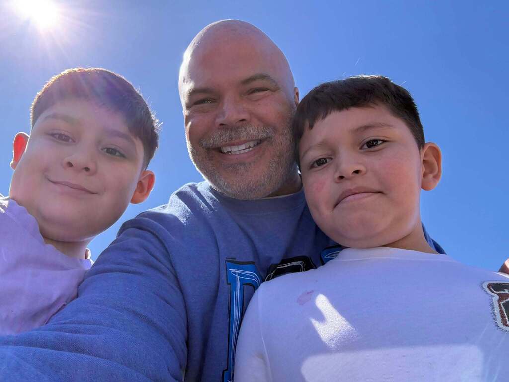 A man and two boys, all smiling, pose for a photo against a bright blue sky.