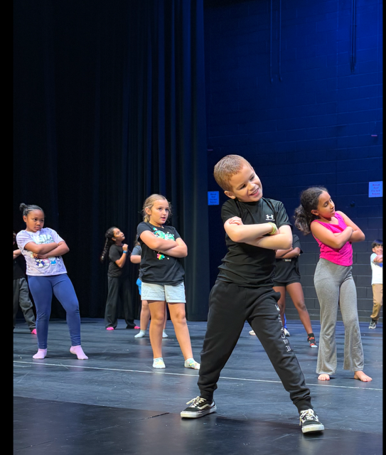 Children in various attire on stage, one boy front center with arms crossed, smiling. Background dark blue.