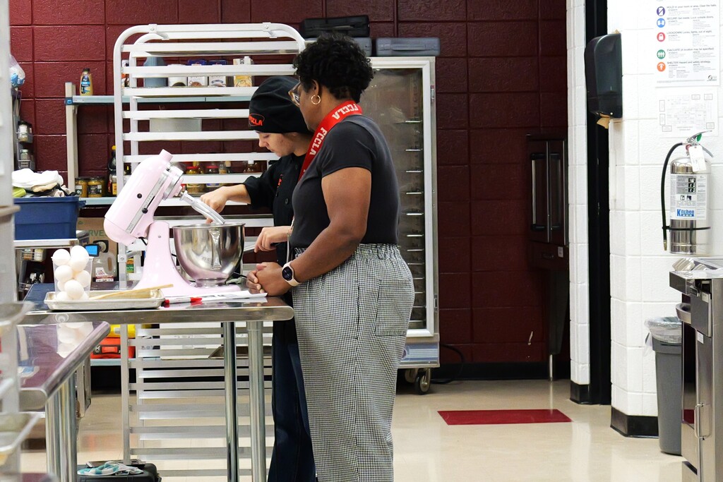 Two people stand in a kitchen, one using a mixer, the other standing by, with equipment and appliances in the background.