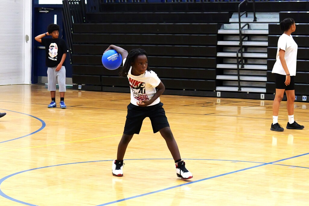 A person dribbles a blue ball on a basketball court. Behind, two people are standing. The court has blue lines.
