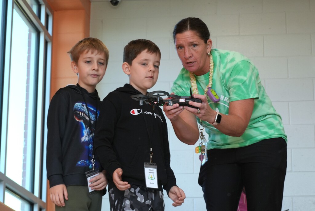 Woman with green top shows two boys a small drone. The boys wear hoodies, one with a dinosaur.