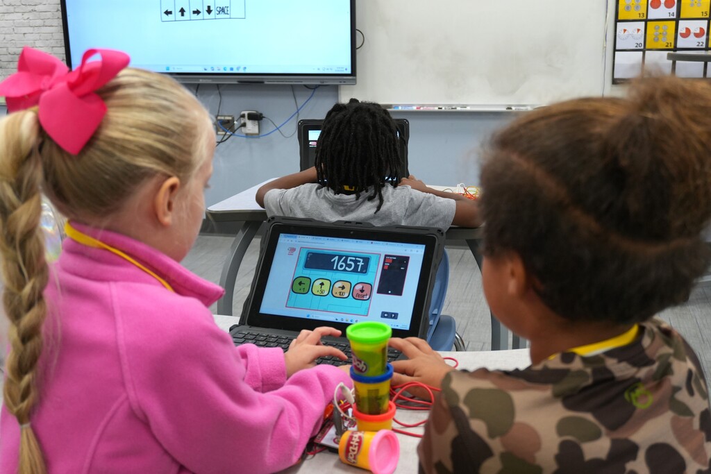 Three children in a classroom, each using a laptop. The girl in pink has a pink bow in her hair.