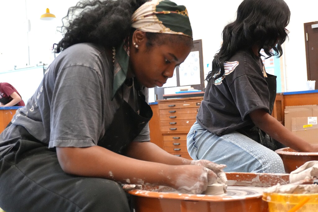 Two women sit in a room, one crafting with clay, the other observing, against a backdrop of cabinets and a partially visible third person.