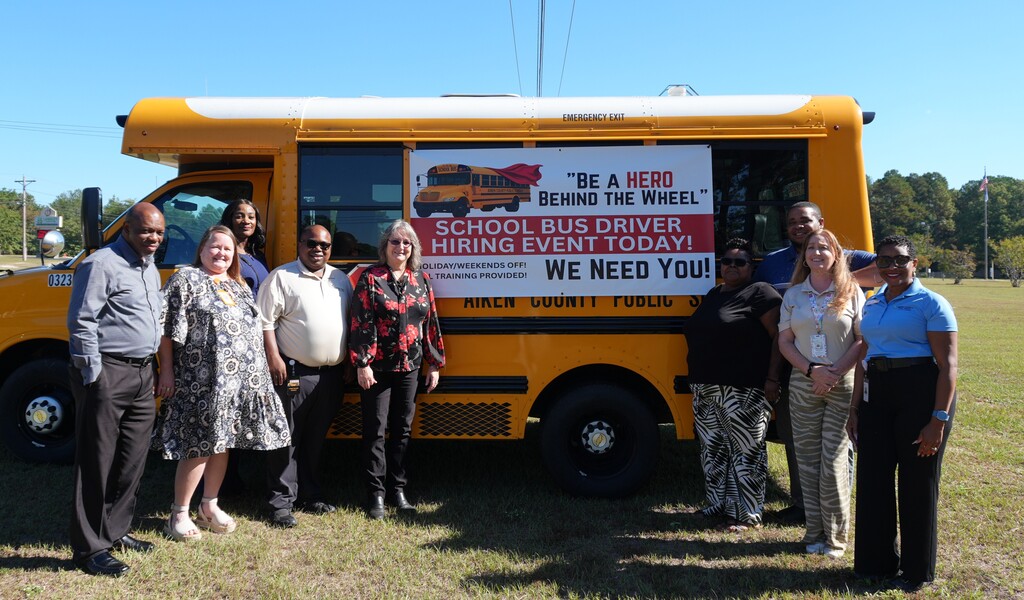 A group of adults stands in front of a yellow school bus with a sign that reads "Be a Hero, School Bus Driver Hiring Event Today. We Need You".