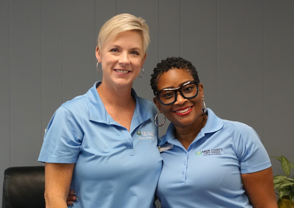 Two women in matching blue shirts pose for a picture with a plant behind them.