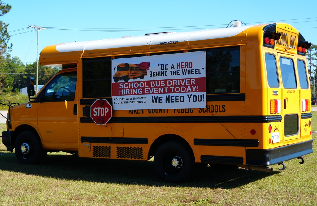 A yellow school bus with a red "STOP" sign parked on grass, displaying a recruitment banner.