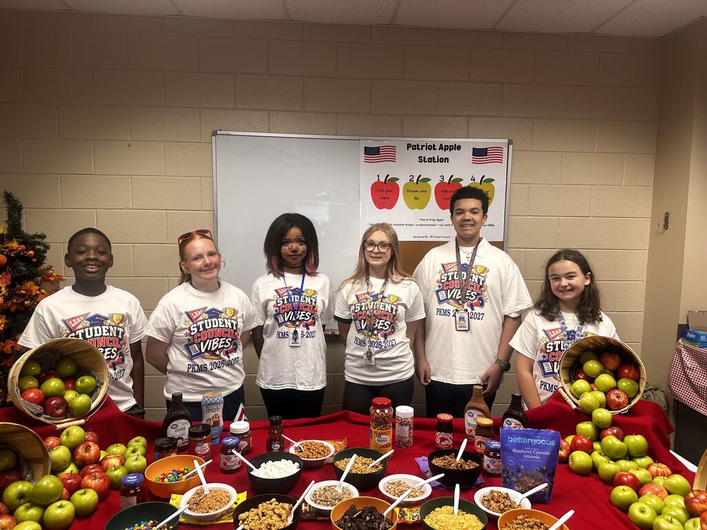 A group of students wearing matching shirts stand behind a table with food. Apples, bowls, and bottles are visible.
