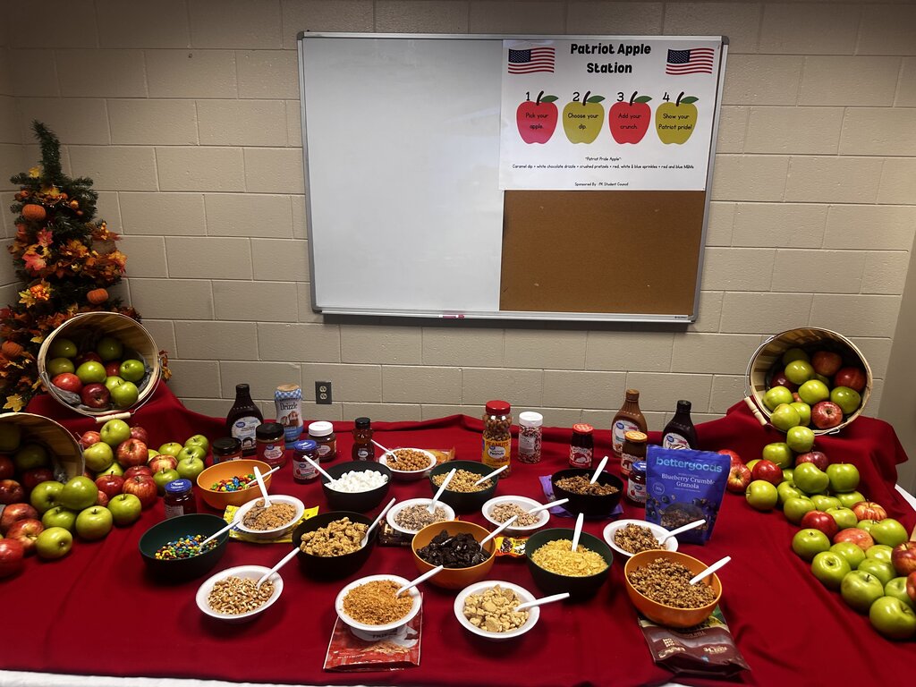 A table with a red cloth displays various dishes, fruits, and condiments. Apples in baskets adorn the background. A whiteboard with an apple design hangs on the wall.