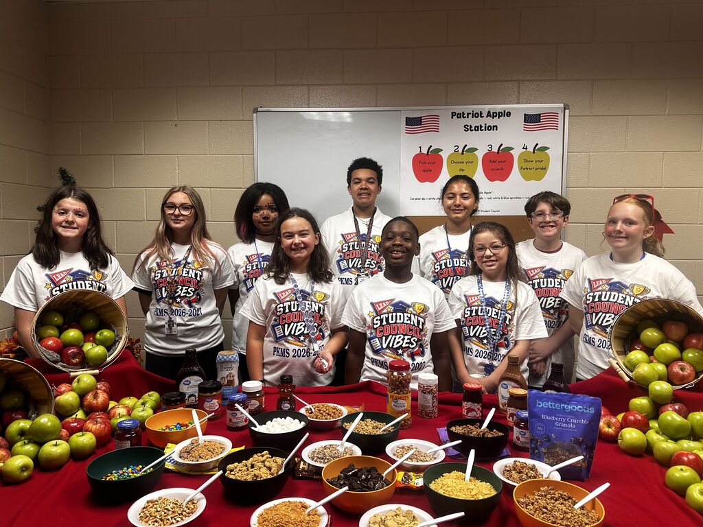 Group of students in matching shirts stand by table with various food, including apples, cereals, and nuts. Behind them, a banner features USA flag and apples.