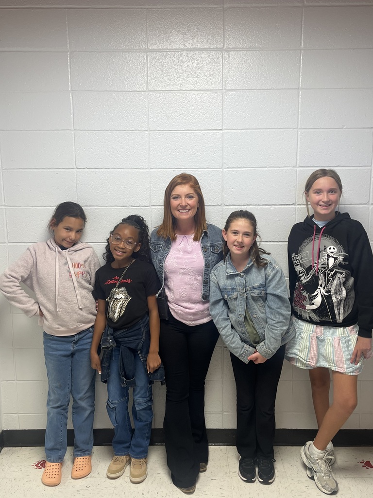 Five individuals stand together against a white wall. Four are young girls in casual clothing. The adult female stands between two girls, smiling.