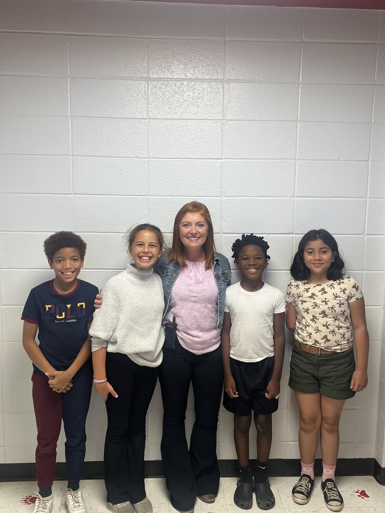 A group of children and a woman standing together. They are smiling. The room has white tiled walls.