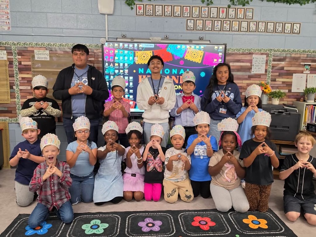 Kids in hats pose for a photo in a classroom. A teacher and students are in front of a screen.