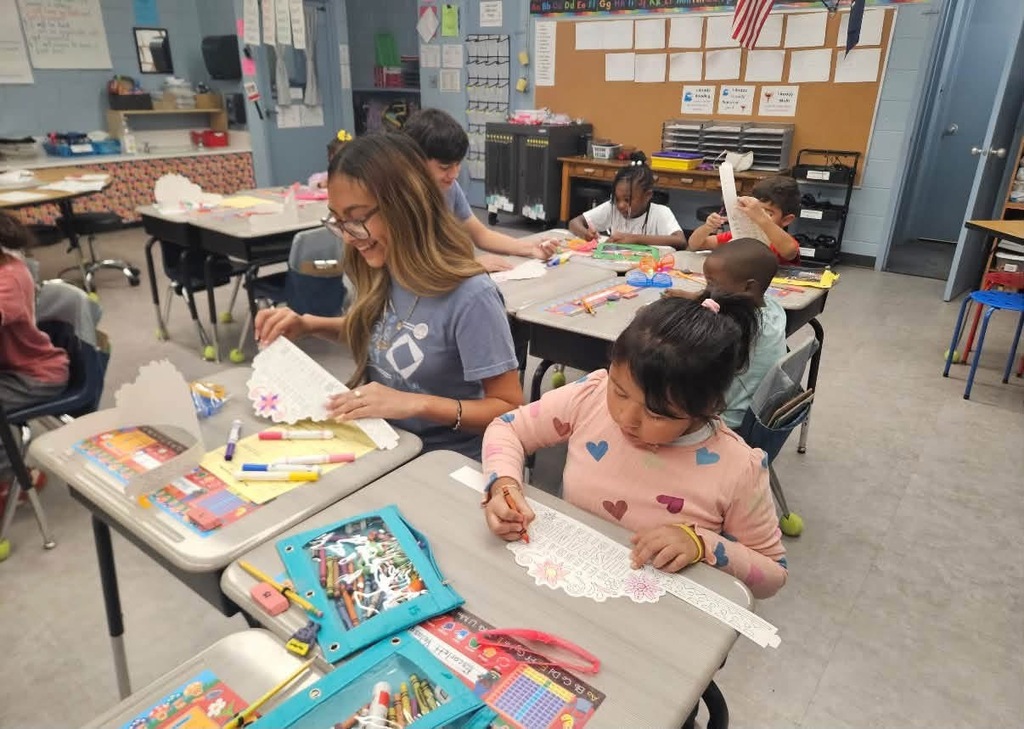 A classroom with students seated at desks. A woman in a blue shirt assists a girl in pink.