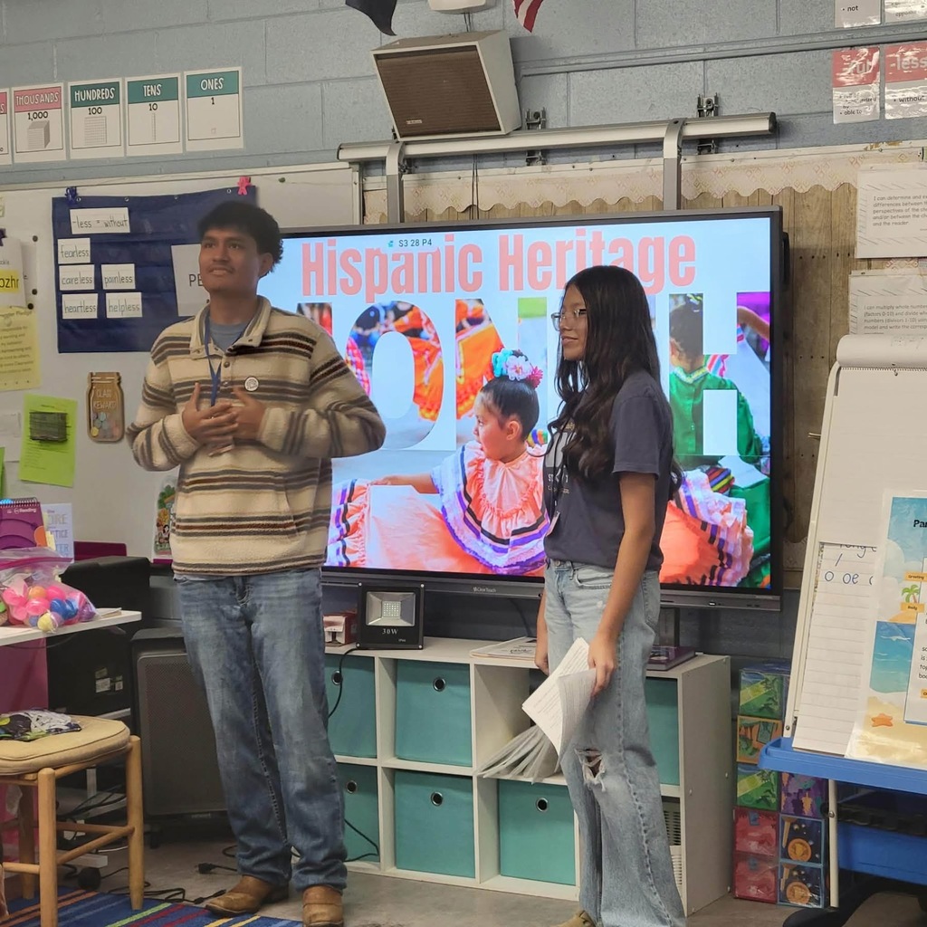 A classroom with a large screen showing Hispanic Heritage. A man and woman stand before it.