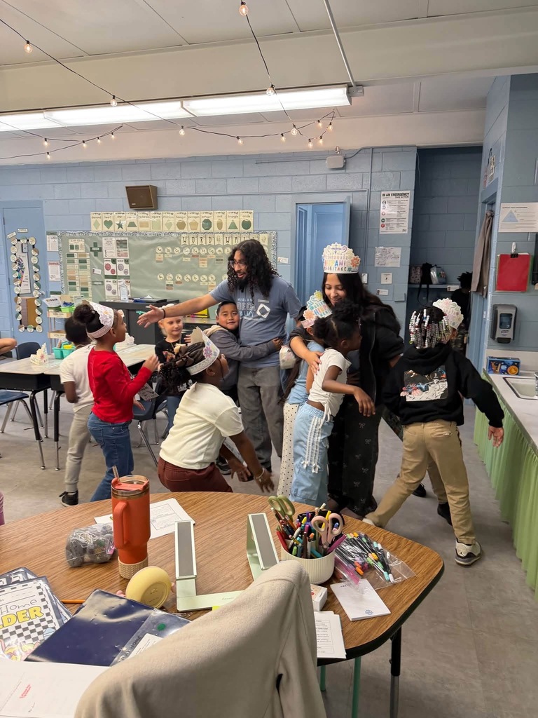 In a classroom, several students and two adults are standing and talking. Some wear headpieces. Desks are set up, and some hold art supplies.