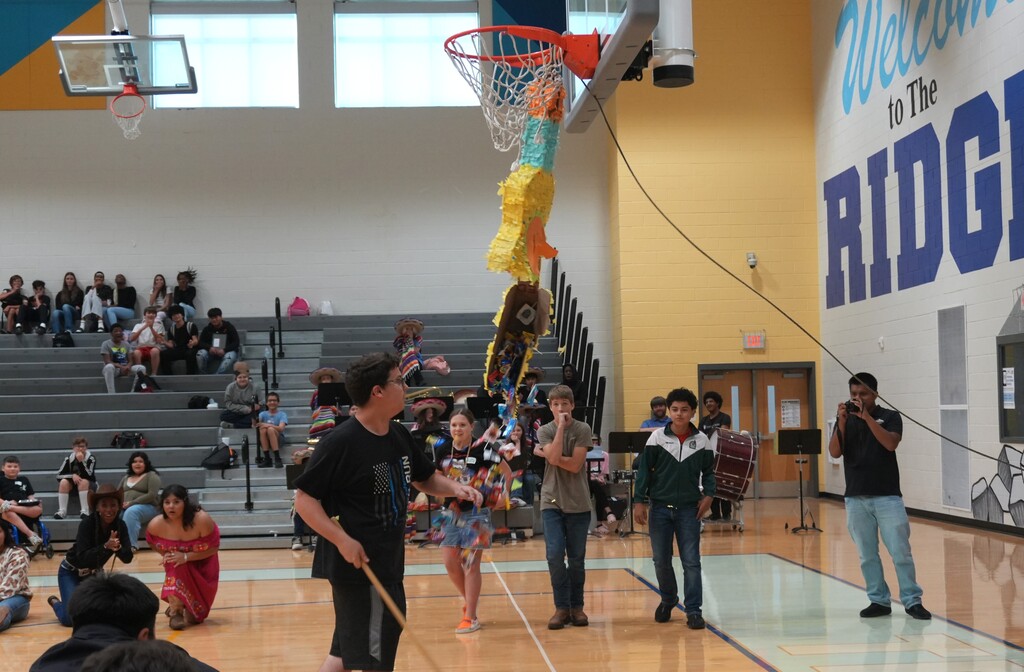 A group of people on a basketball court. Some are performing while others watch. A colorful object hangs from the hoop.