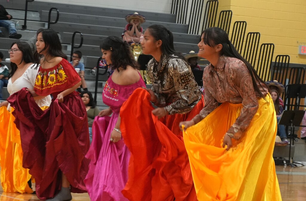 Five women in colorful dresses perform a dance routine on a basketball court. Behind them, people sit on bleachers.