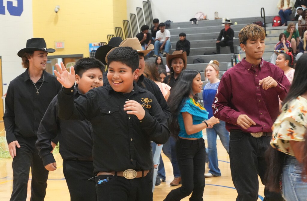 Several people dancing in a gymnasium, one with a cowboy hat and wide smile, surrounded by others.