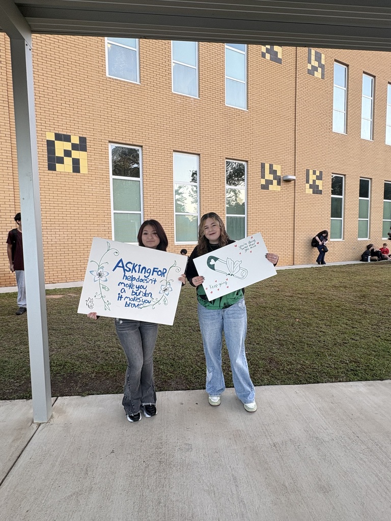 Two individuals, each holding a sign, stand in front of a building with glass windows. A grassy area and additional people are behind them.