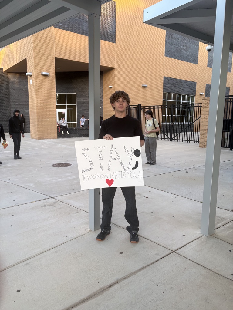 Student holding sign in front of school with some students in the background. The sign says "STAY" and is hand drawn. 