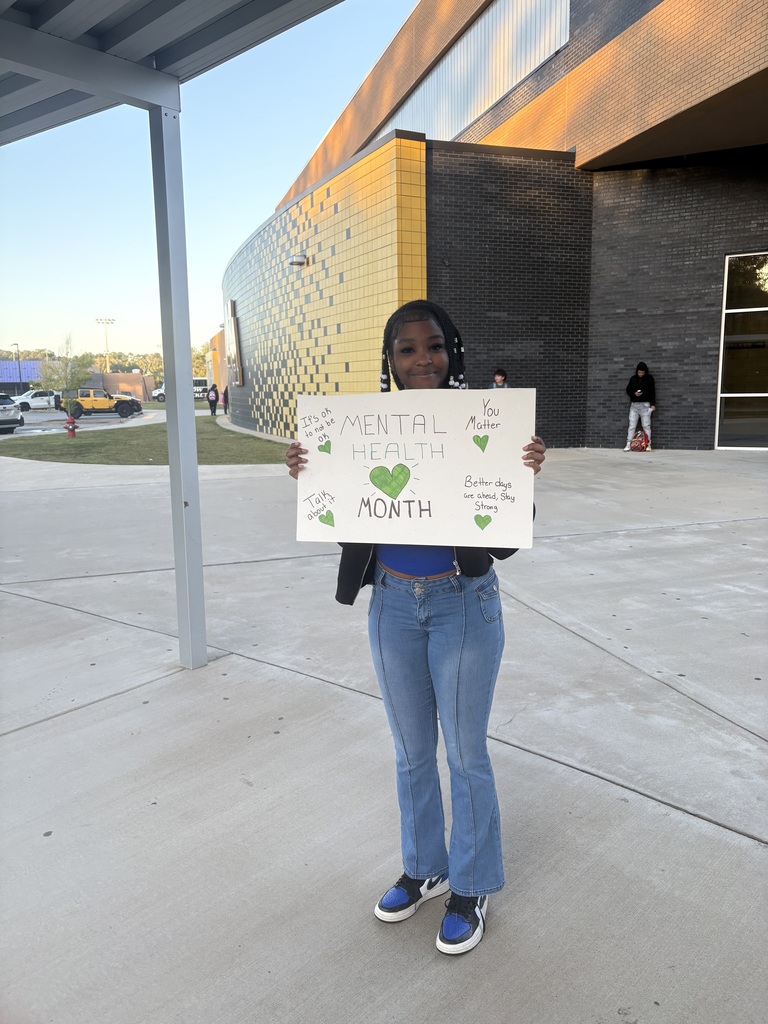 Student standing in front of scho9ol with a hand drawn sign that says "Mental Health Month"
