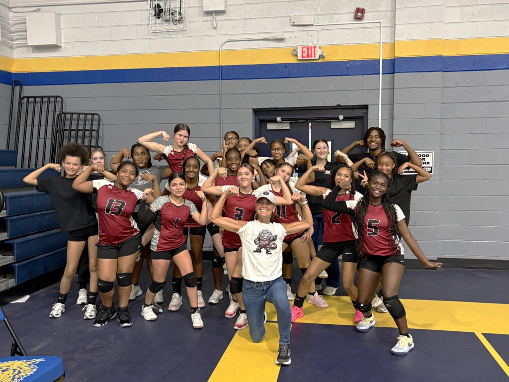 A group of women volleyball players posing with coach in a gym. Yellow and blue floor.