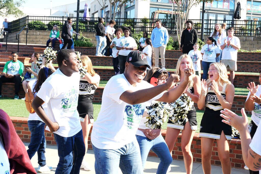 People dance with pom-poms in front of seated spectators at an outdoor event.