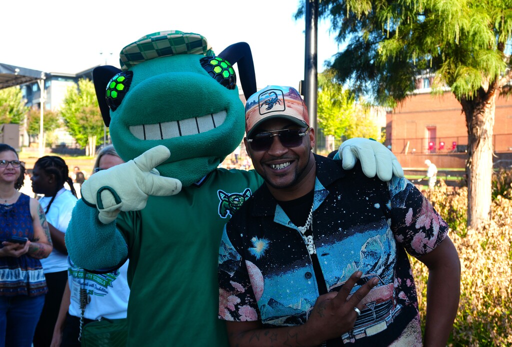 A man in a floral shirt and cap poses with a mascot dressed in a green costume.