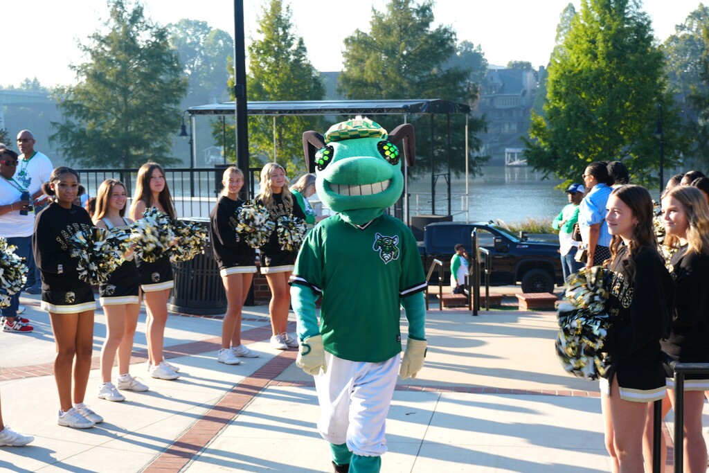 People standing in line, including cheerleaders, a mascot, and others, on a sidewalk with trees and buildings.