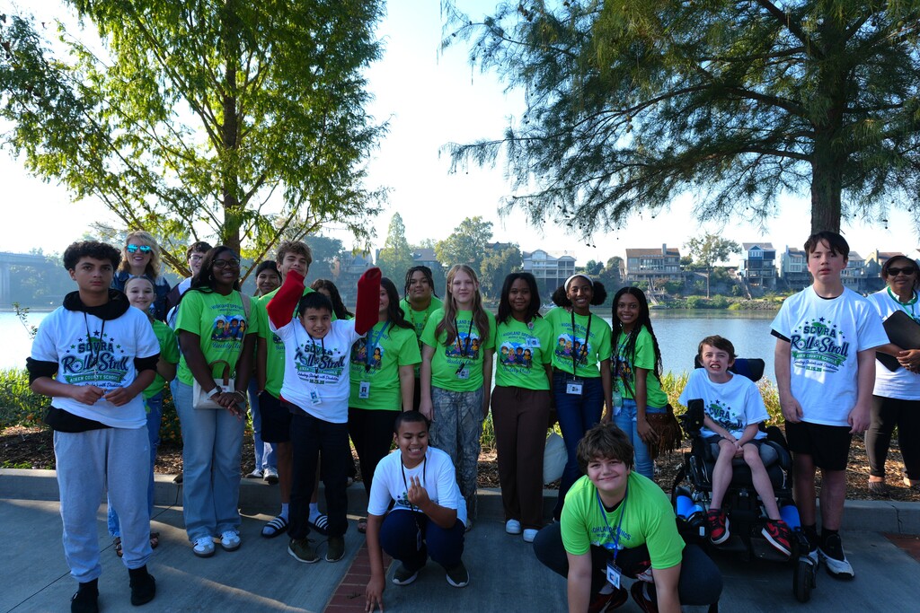 Group of students in green shirts stand and crouch in a line outside by a lake.