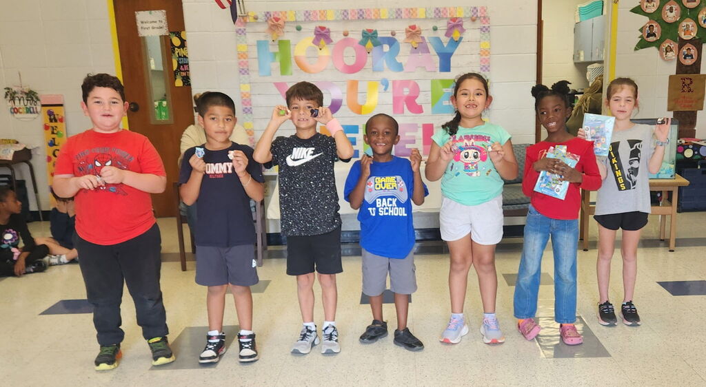 Nine children in a classroom stand in a line, showing off badges and books. A banner reading "HAPPY SUMMER" hangs behind them.