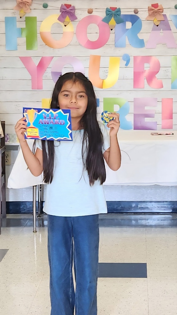 A girl holding a certificate stands in a room with a colorful banner reading "Hooray You're".