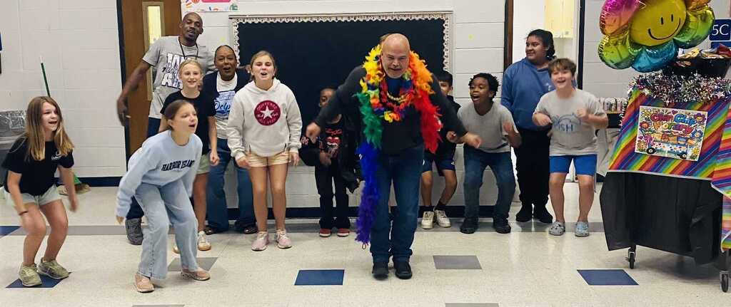A group of students, teachers, and a man wearing a rainbow scarf pose for a photo.