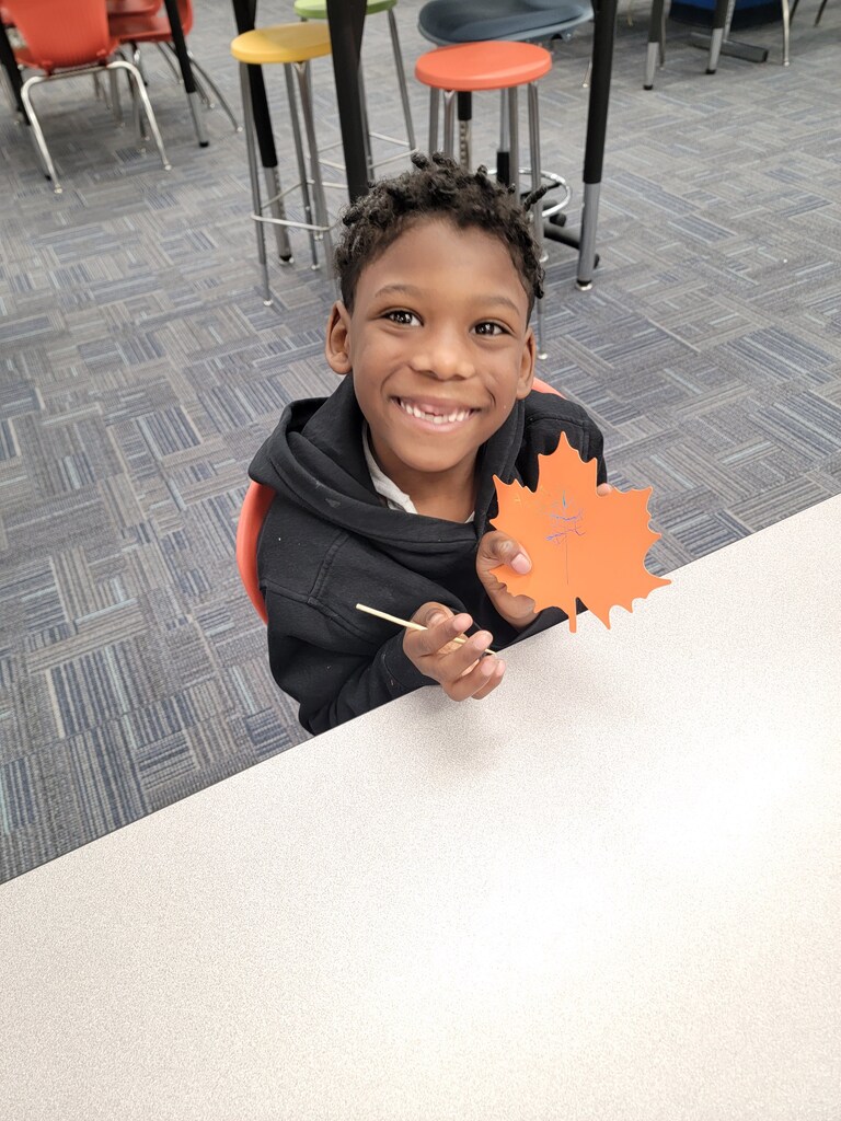 Young boy holding a leaf, smiling at the camera, sitting on a chair in a classroom.