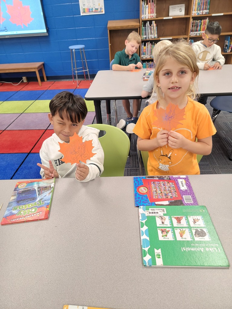 Two children seated at a table, one holding a leaf, the other holding a book.