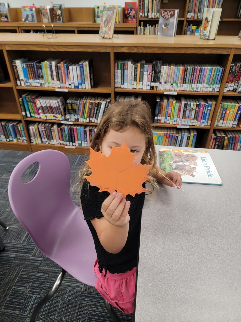 A young girl sits in a purple chair holding up an orange leaf in front of her face.