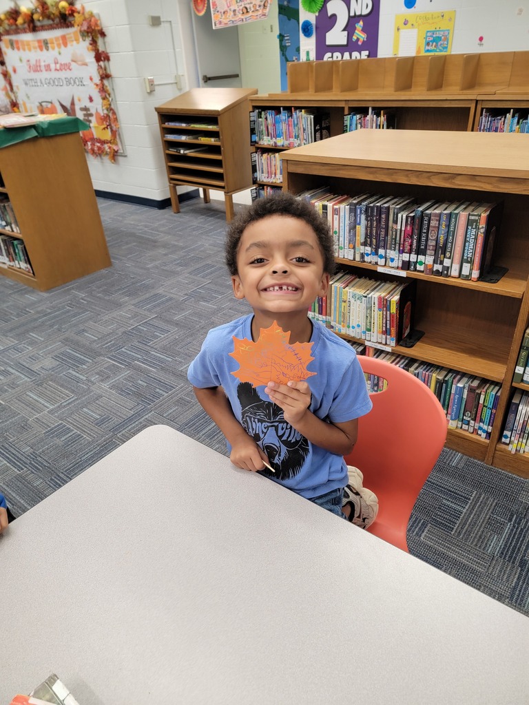 A child smiles while holding a leaf, seated on a red chair at a table in a library.