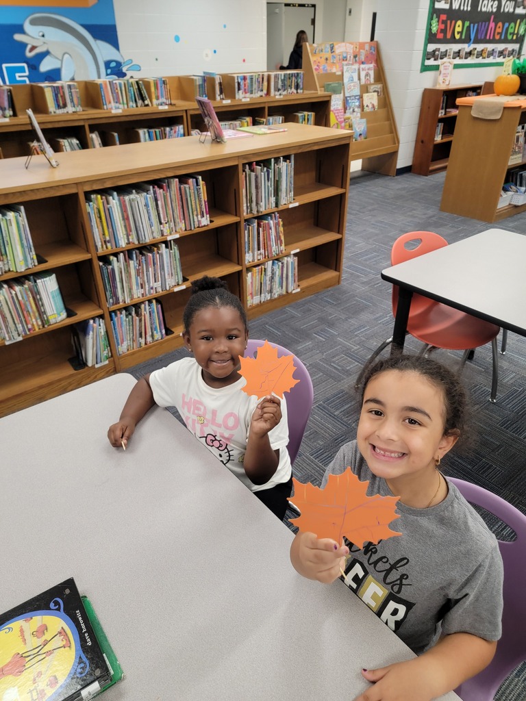 Two children at a table, one holding a red leaf, sitting in chairs with books on shelves behind them.