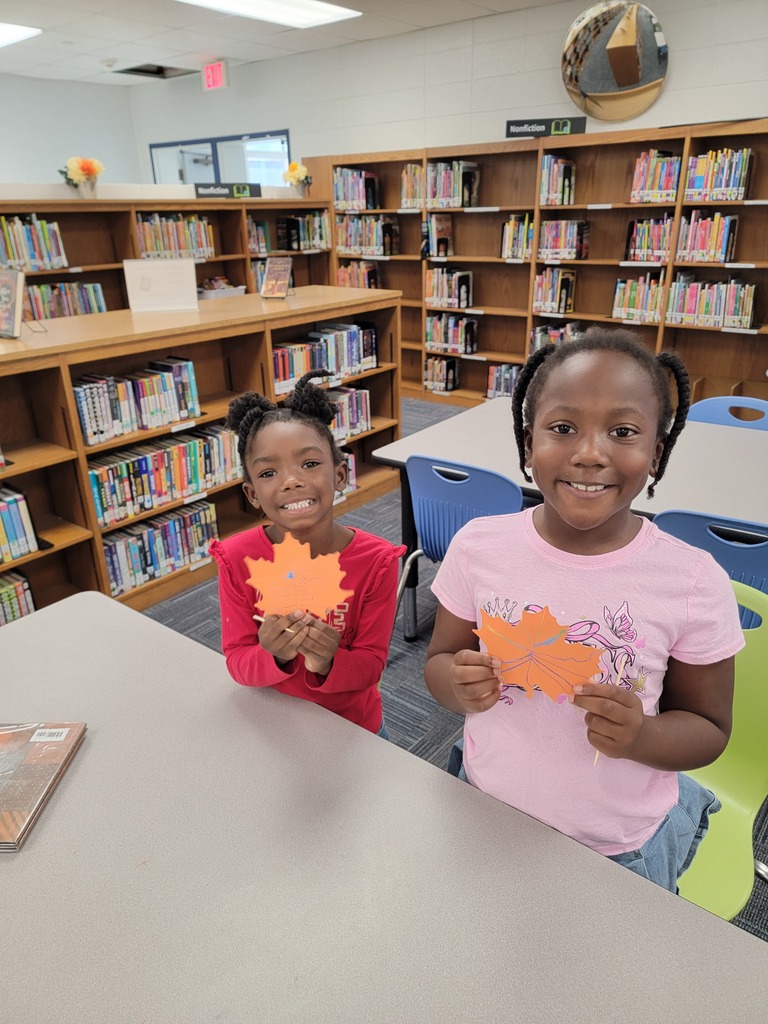 Two smiling children holding orange paper cutouts, standing in front of a library shelf.