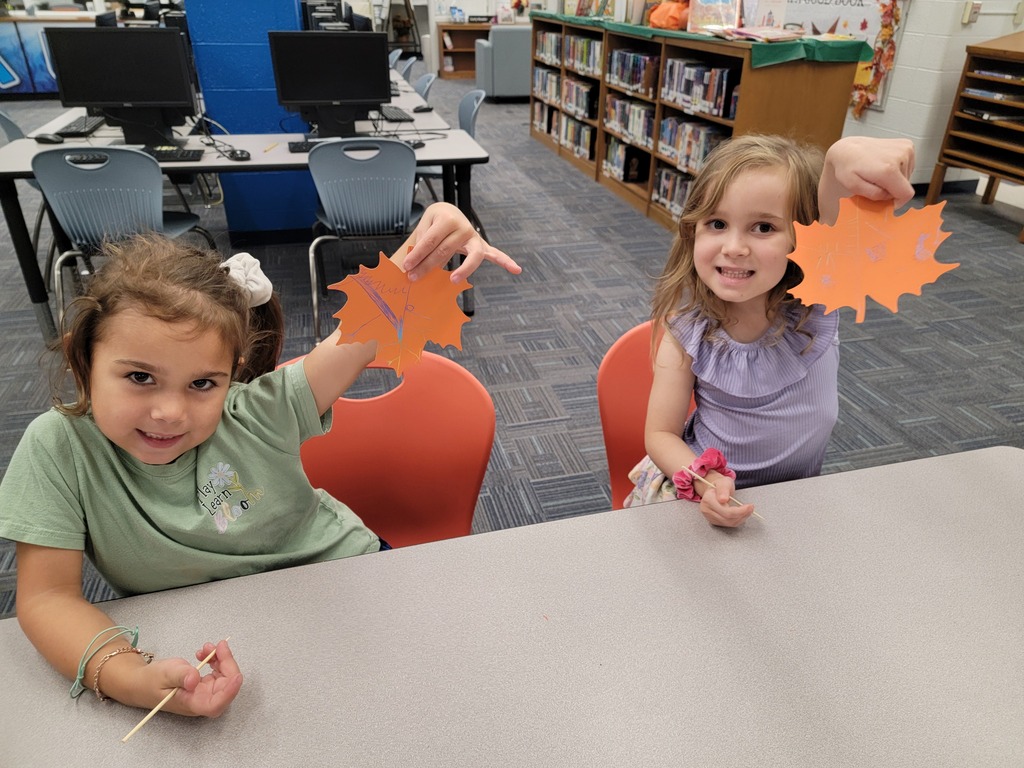 Two children holding orange leaves, seated at a table in a library with bookshelves and computers.