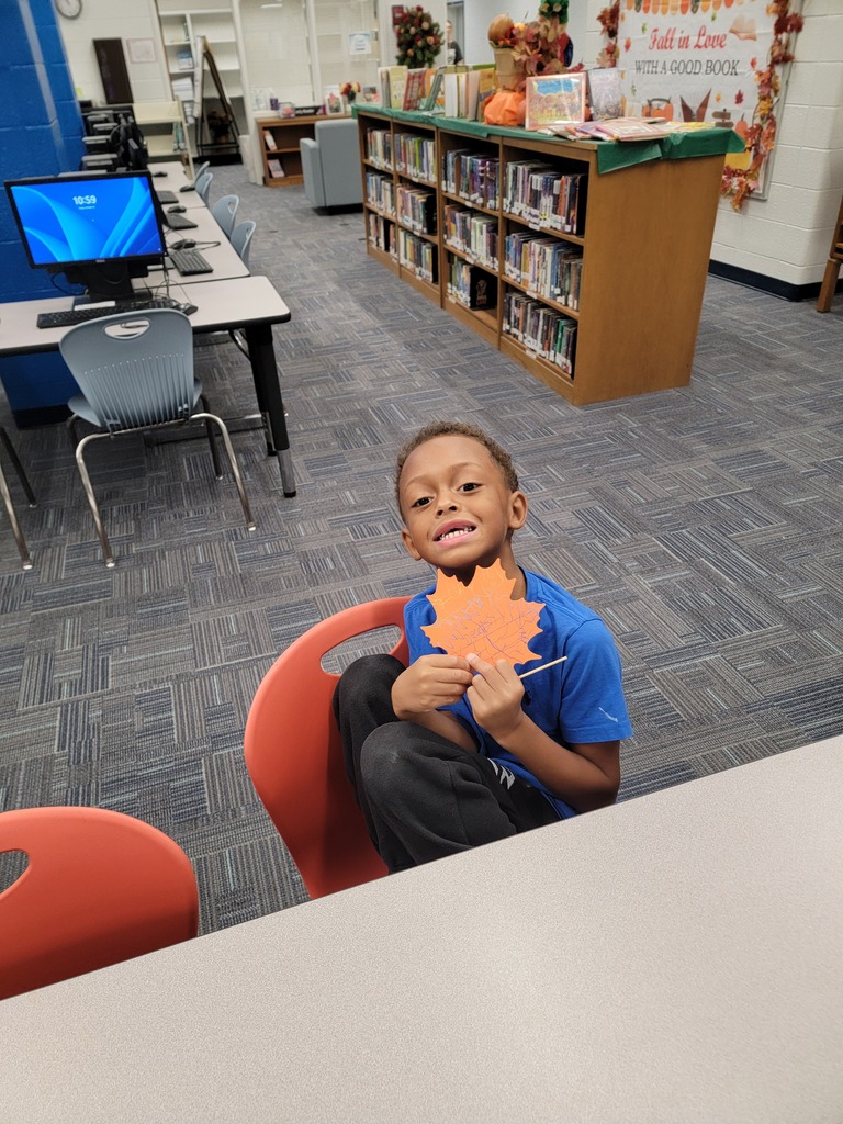Child in blue shirt holding a leaf-shaped paper sitting on a red chair in a classroom.