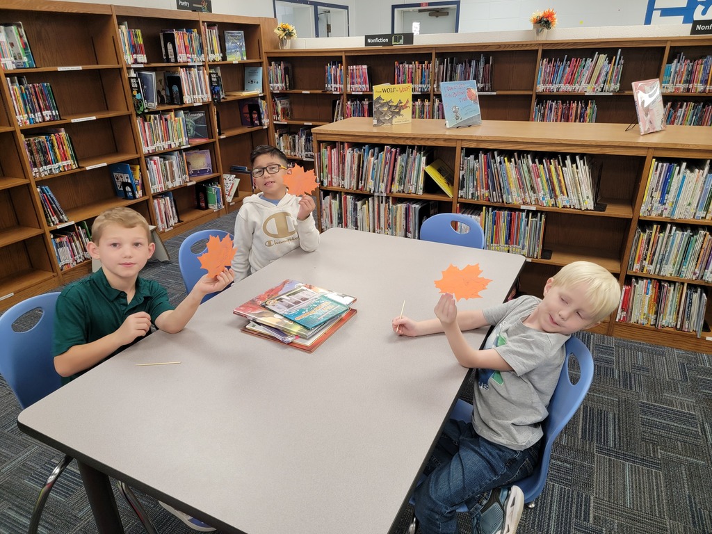 Three children sit at a table in a library with orange papers, one holding a book.