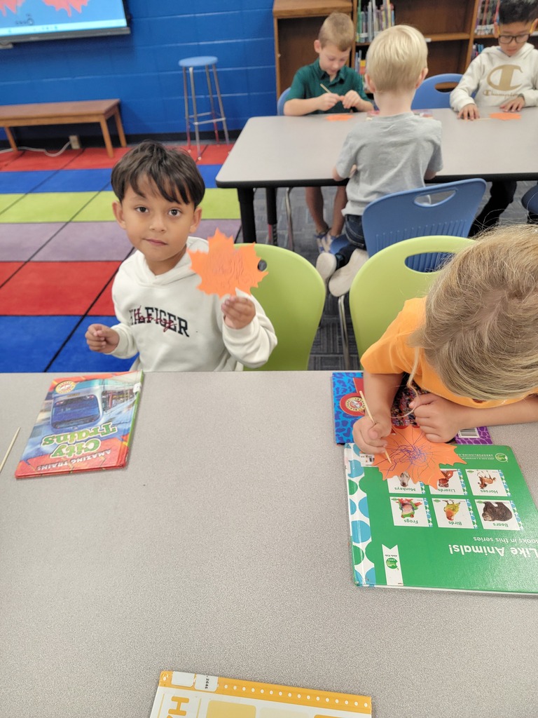 Children seated at a table, one holding a leaf, surrounded by books, a multicolored wall, and a bookshelf.