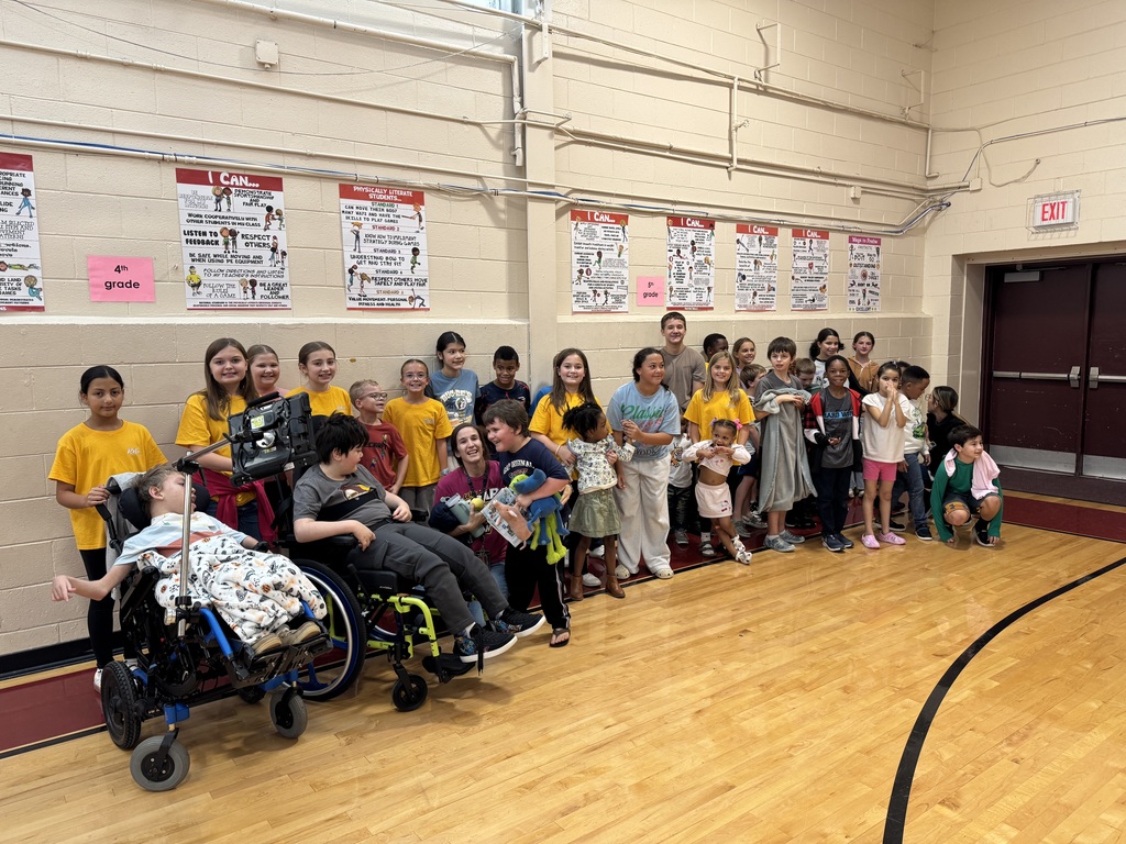 Students in yellow shirts gather in a gym; some sit in wheelchairs, others stand. A black line marks the court.