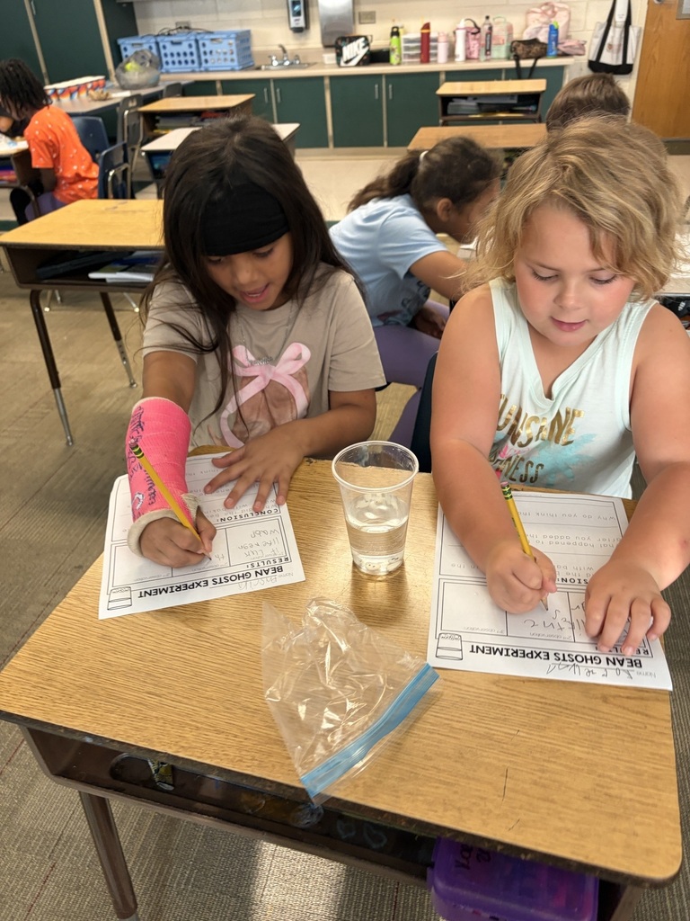 Children seated at desks write on papers; one child has a bandaged arm, another holds a pencil.